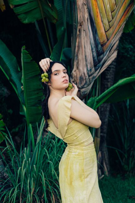 A woman in a yellow dress poses in a tropical environment holding grapes.
