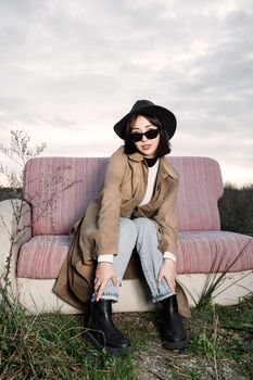Fashionable woman posing on an outdoor sofa wearing a trench coat, jeans, and sunglasses.