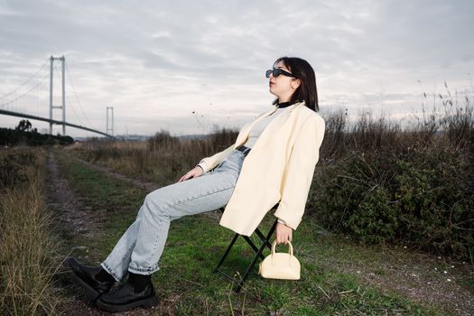 A stylish woman in sunglasses relaxes on a chair by a bridge in nature.