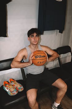 Young man with basketball sits on bench indoors, ready for game.
