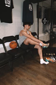 Young man preparing for a basketball game in a gym locker room, lacing up his sneakers next to a basketball.
