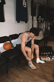 A young man in a gym locker room tying colorful basketball shoes next to basketball gear.