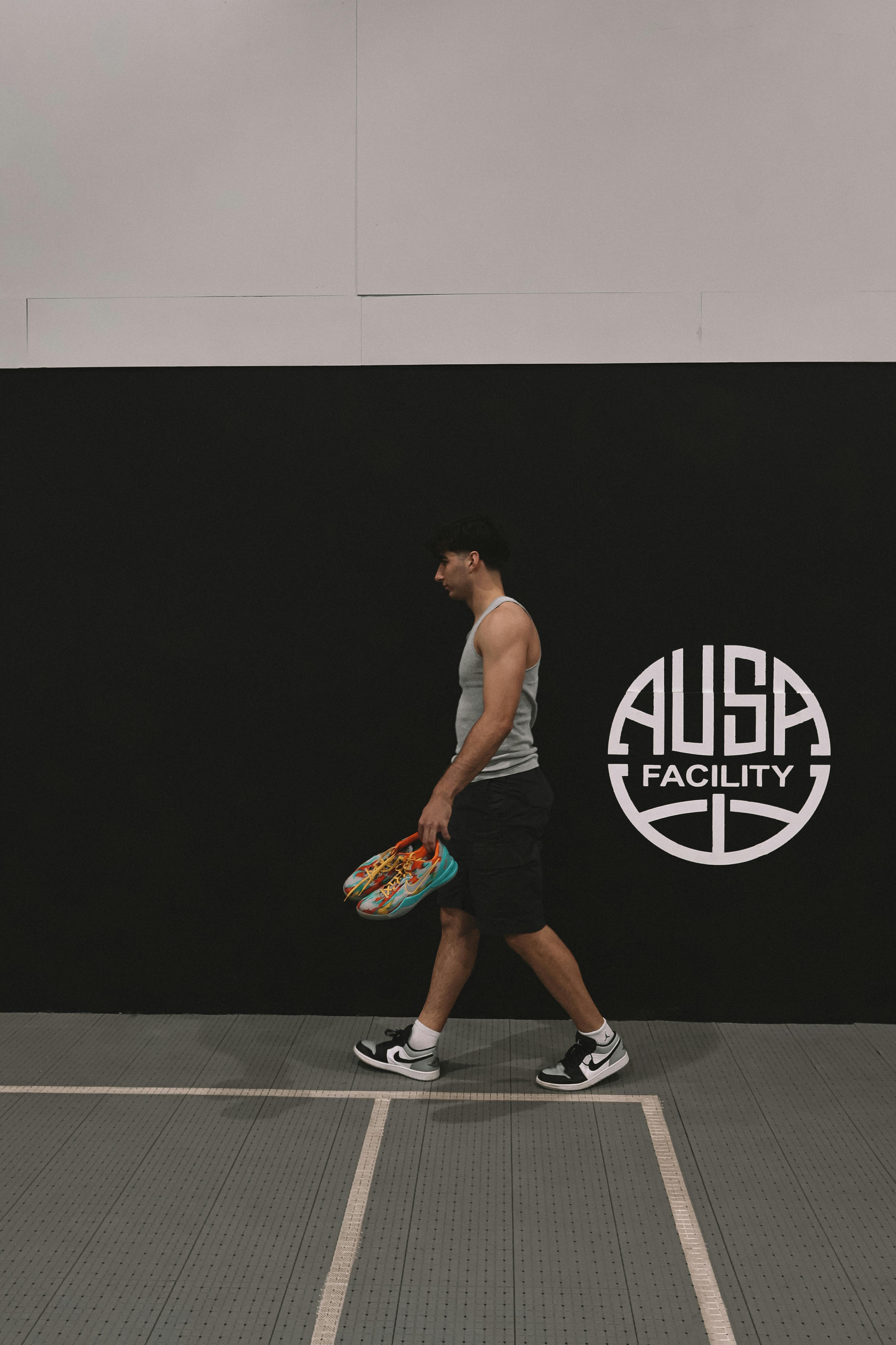 Man walking in gym with colorful ball near AUSA Facility wall, wearing sporty outfit.