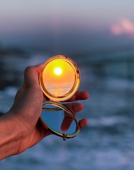 Hand holds mirror reflecting a vibrant sunset on a scenic beach.