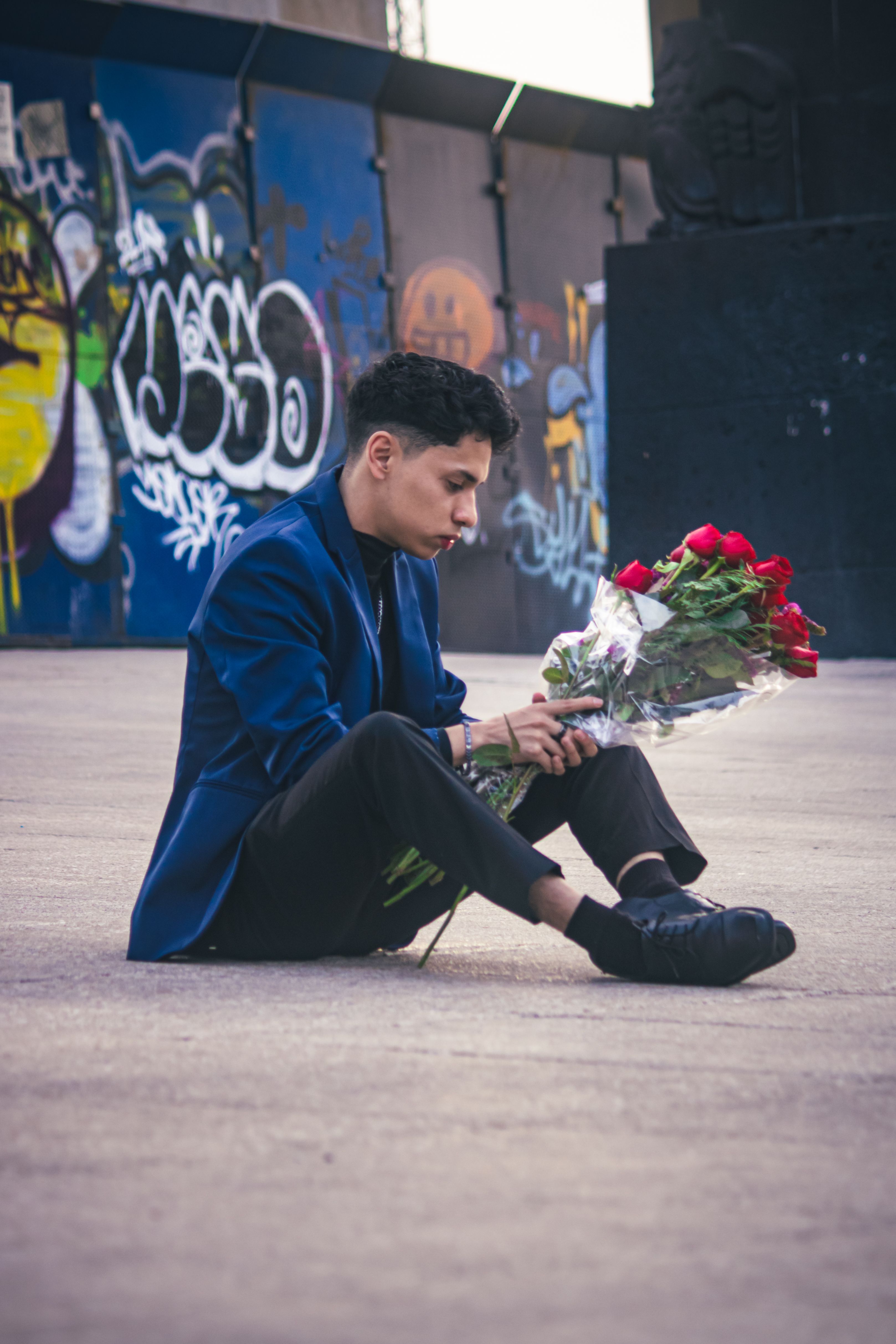 Young man in blue jacket sitting with roses in a graffiti-covered urban area.