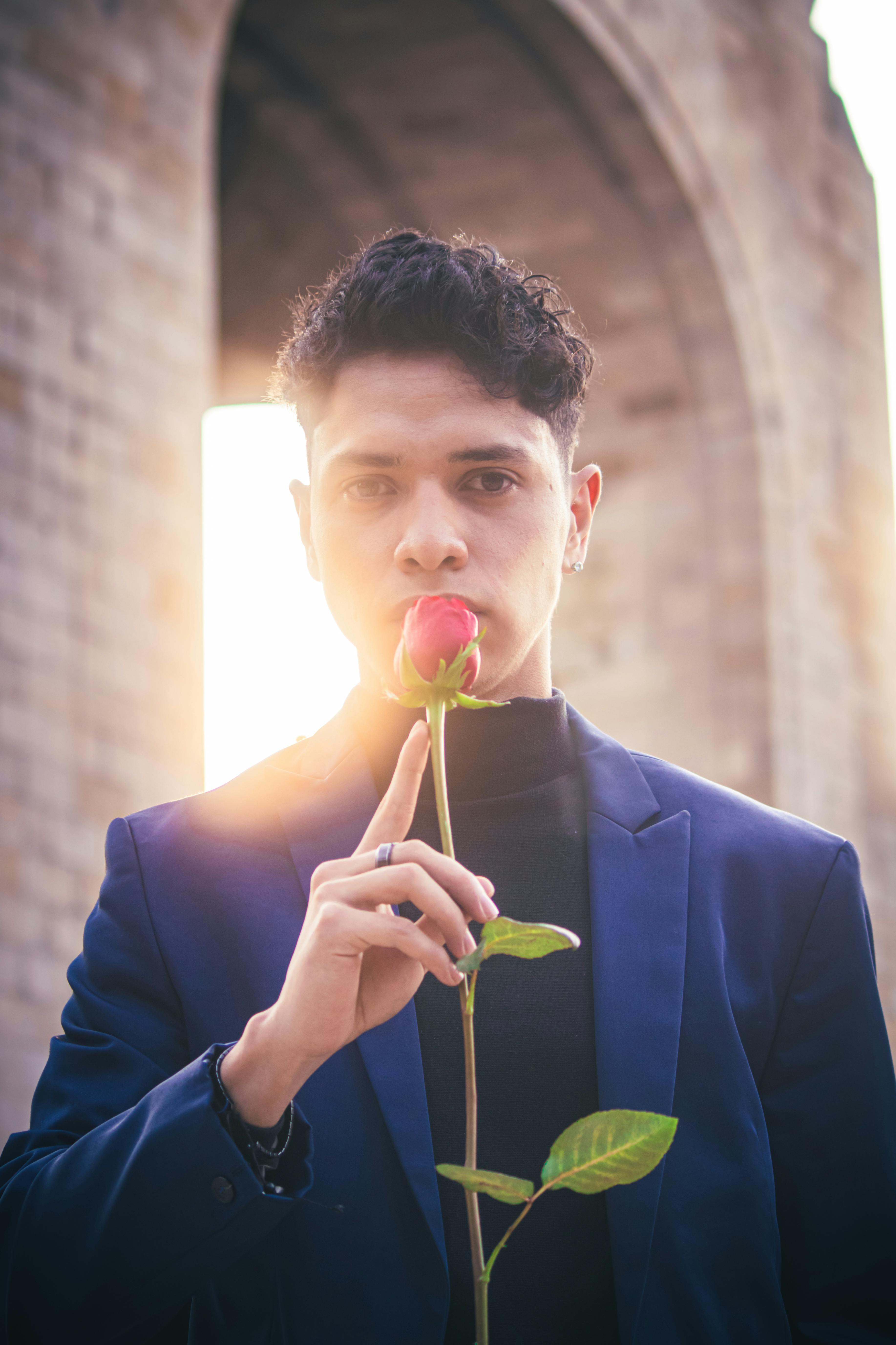 Young man holding a red rose in front of a historic archway at sunset, capturing a romantic vibe.