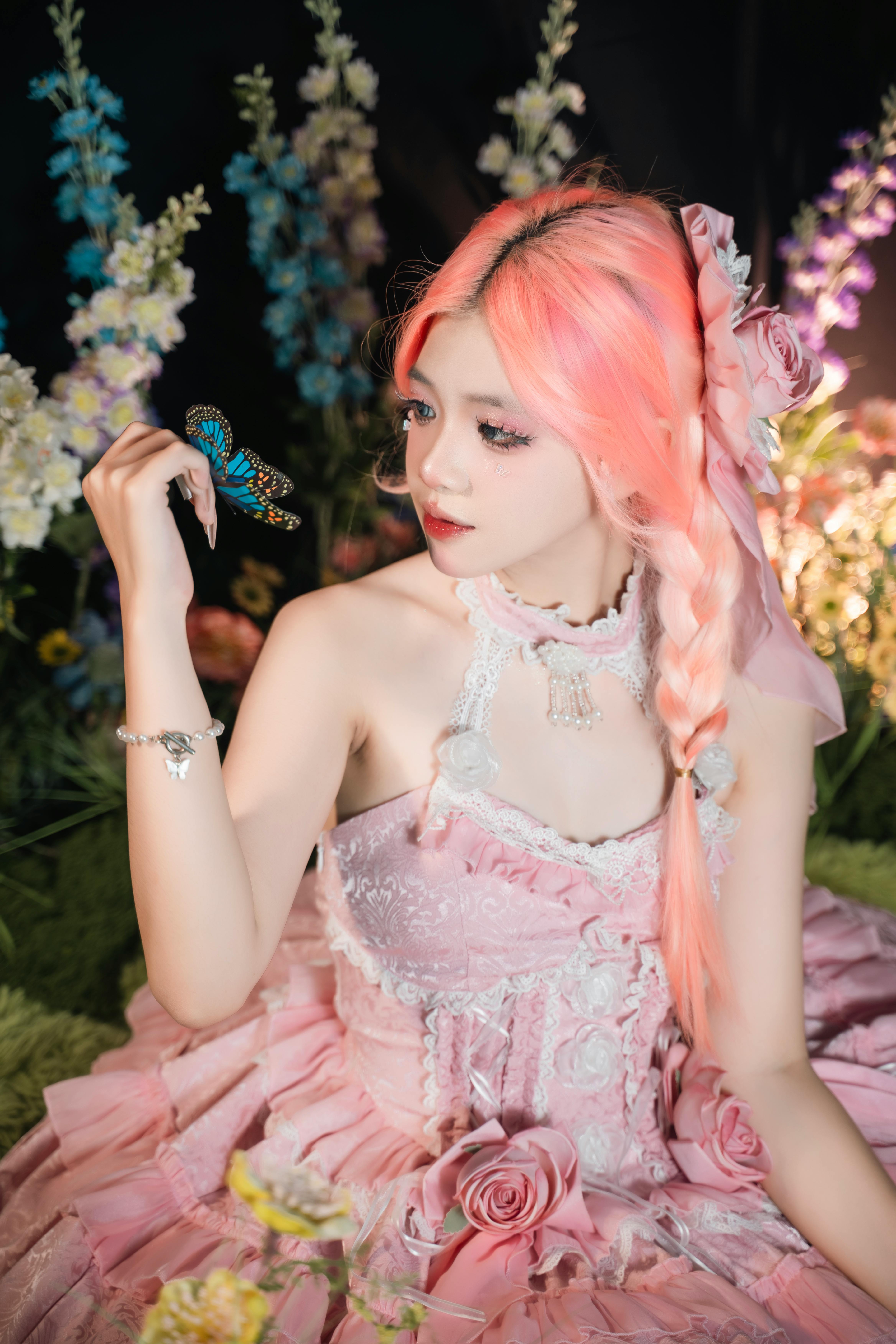 A woman in a pink dress with pink hair, holding a butterfly surrounded by flowers.