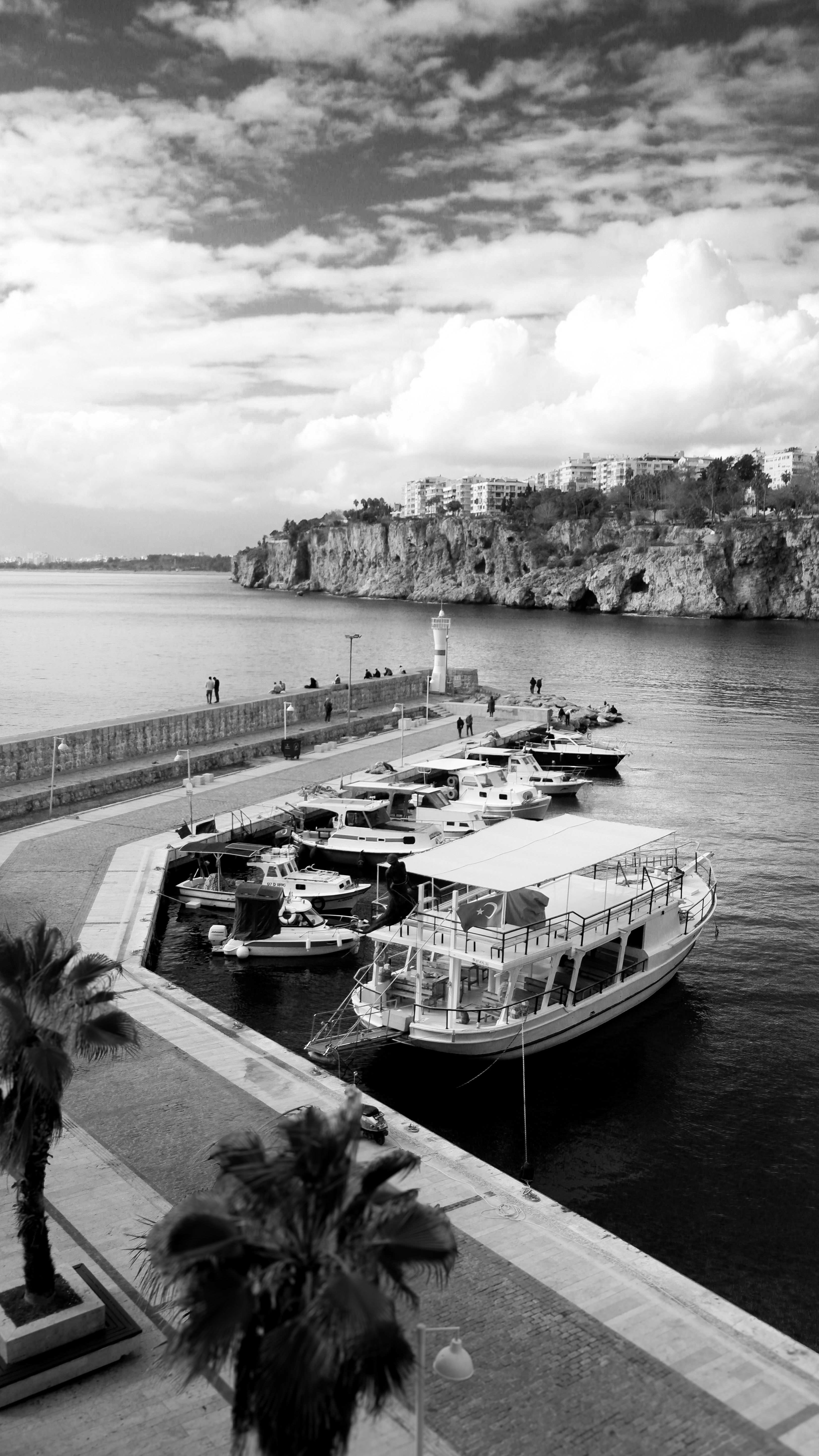 Black and white photo of a harbor with yachts and cliffs under a dramatic sky.
