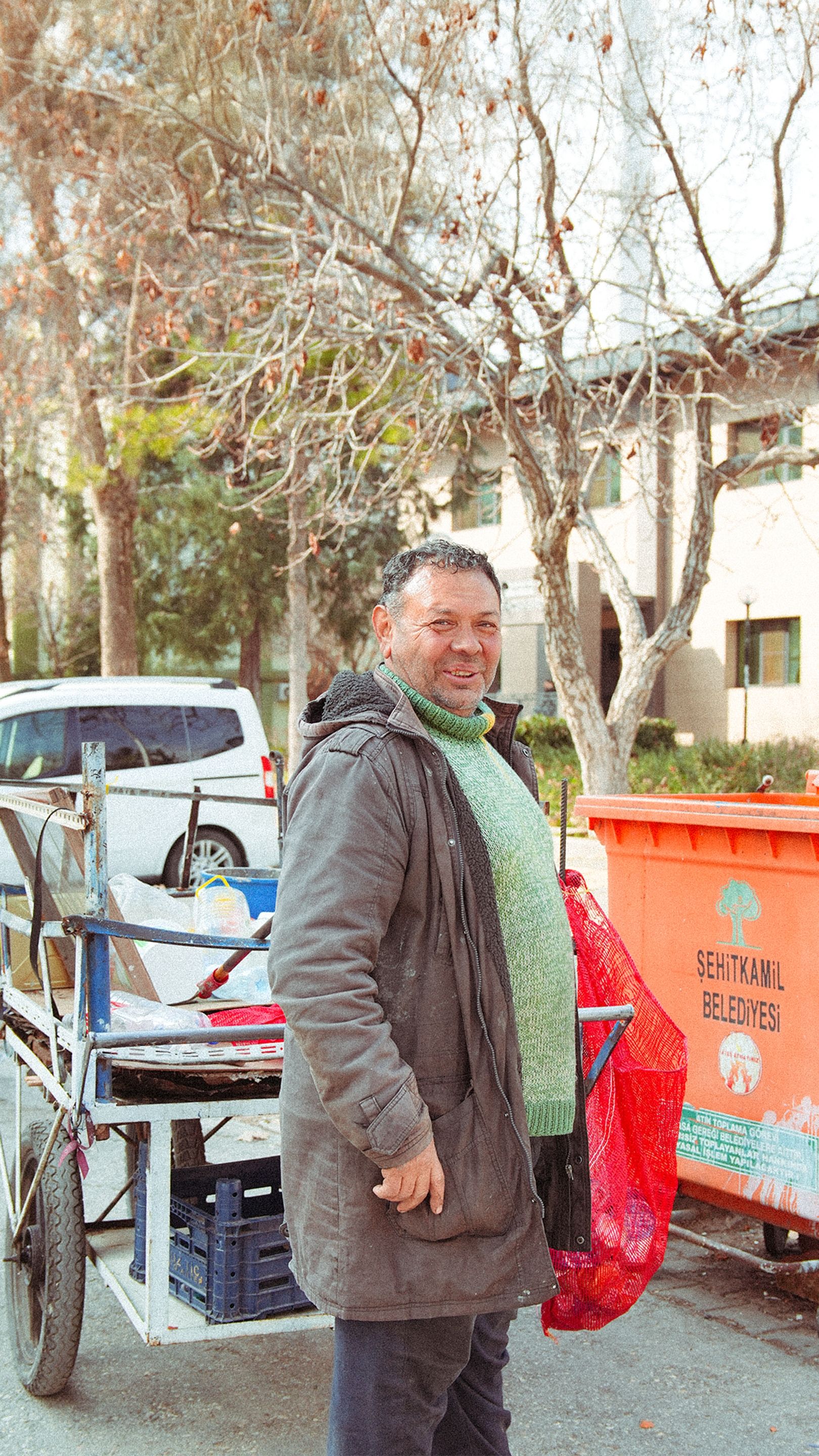 A man stands next to a barrow on a tree-lined street in Gaziantep during daytime.