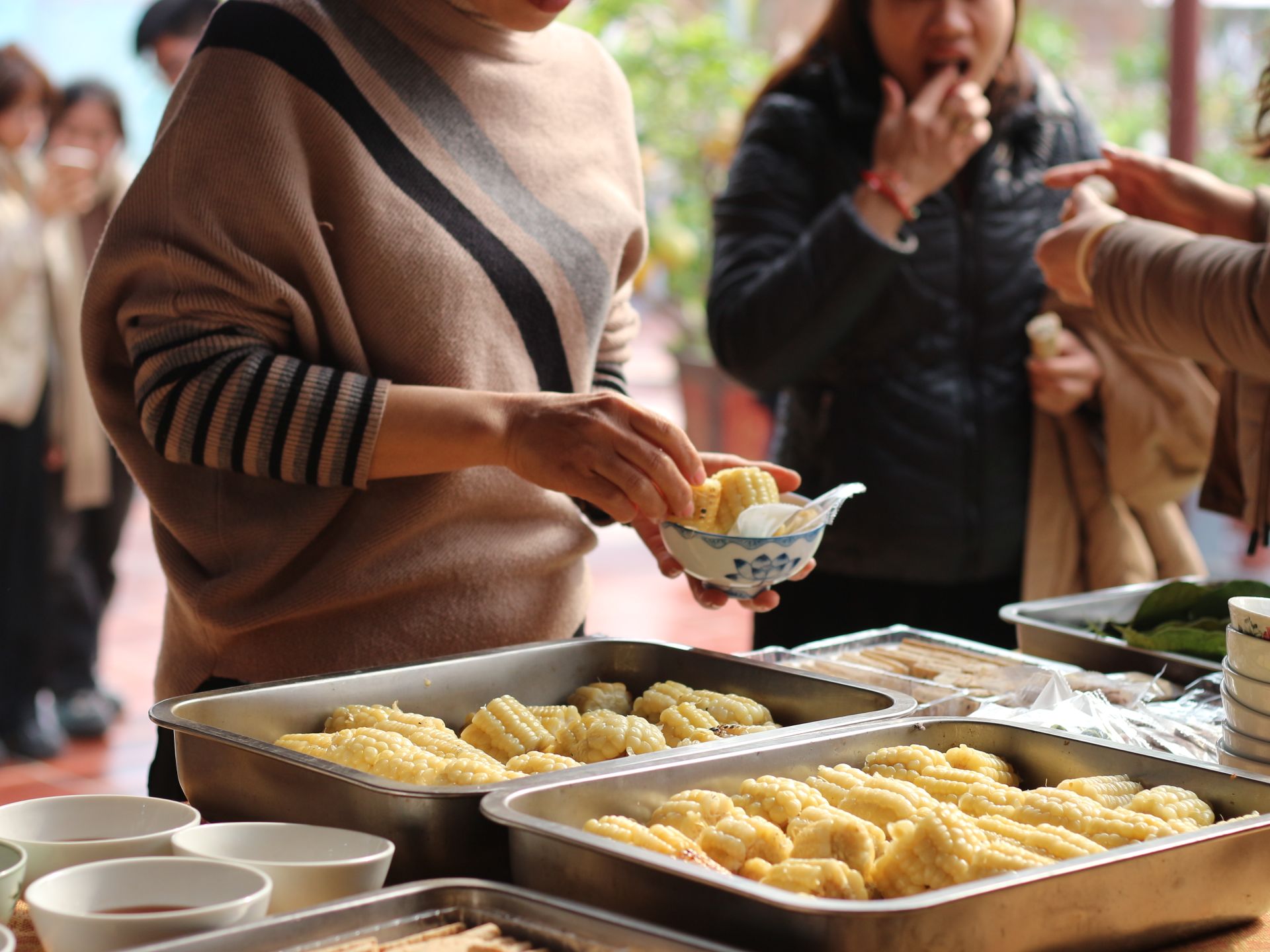 A woman serves corn pieces at an outdoor food stall with people around.
