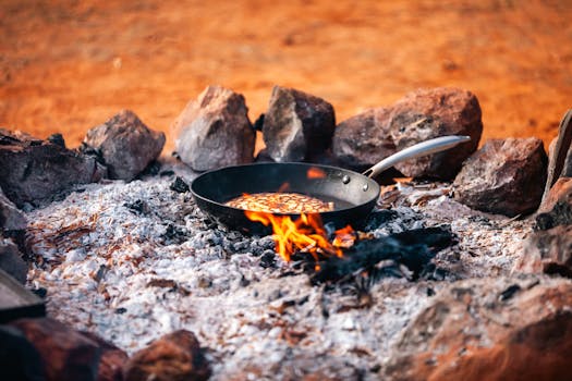 A rustic scene of cooking pancakes over a campfire using an iron skillet surrounded by stones.