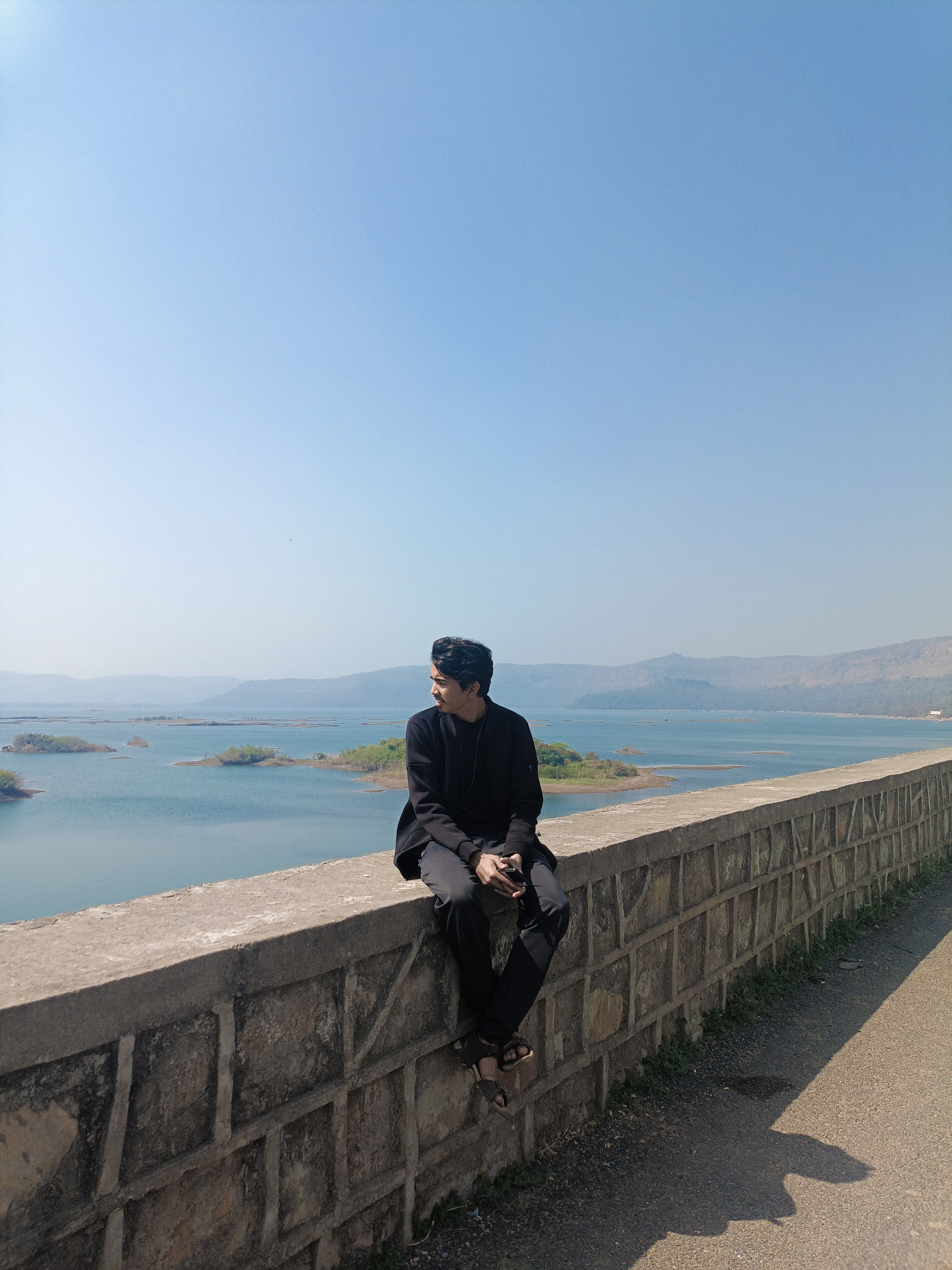 A man in a black outfit sits on a stone wall, enjoying the tranquil view of a lake and mountains.