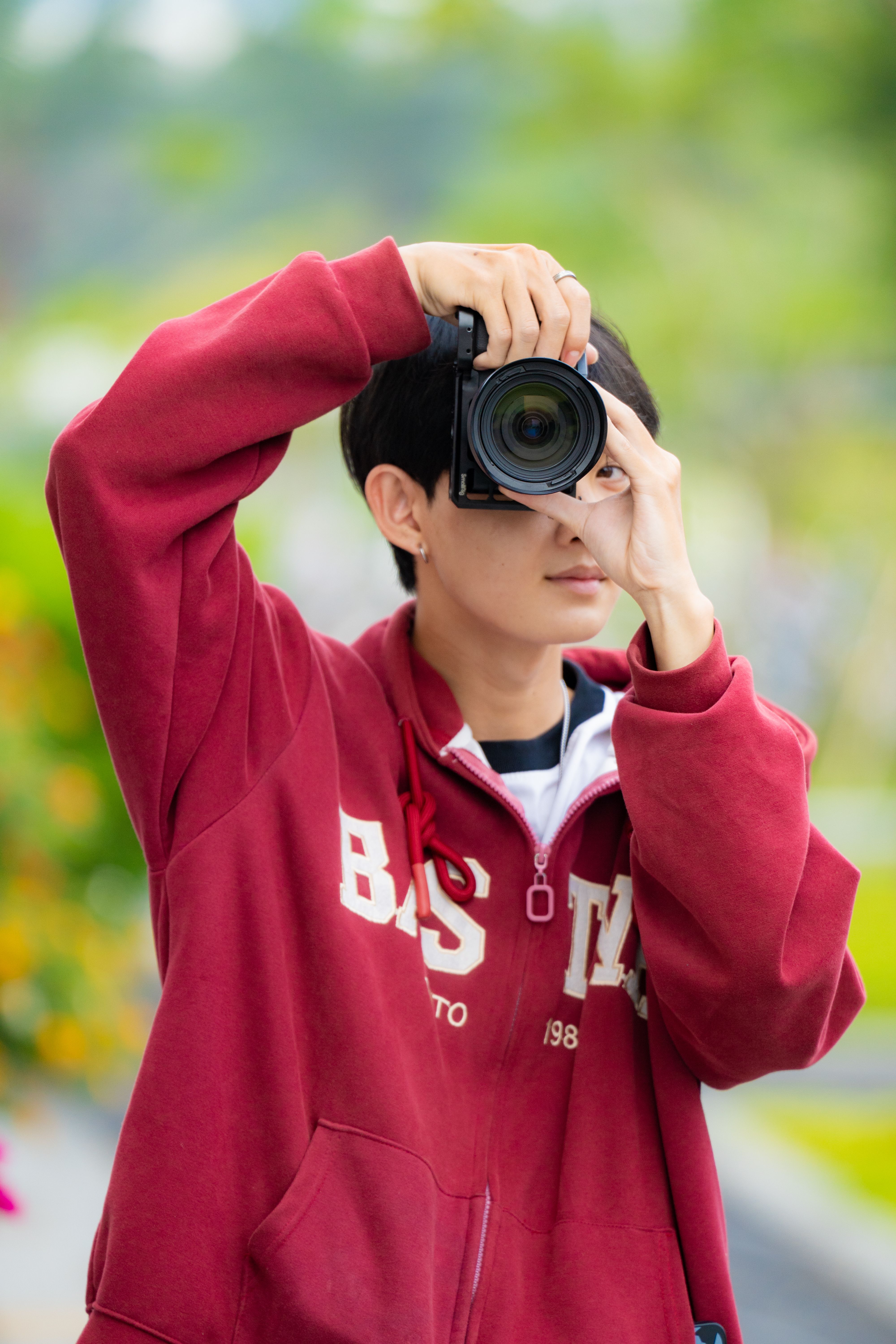 A person wearing a red jacket taking a photo with greenery in the background.