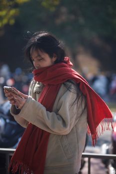 A young woman in a red scarf texts on her phone outdoors, capturing a serene winter moment.