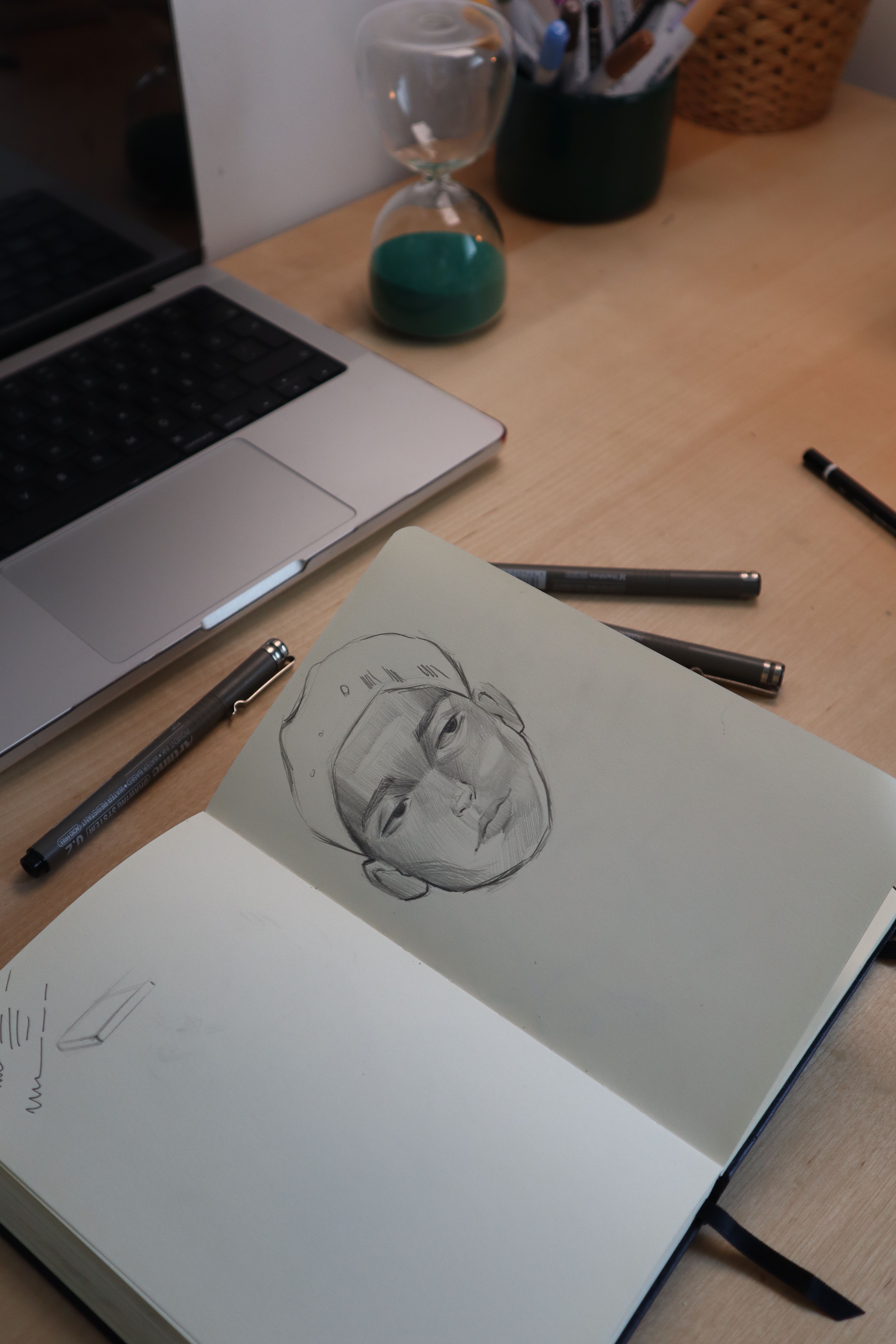 Creative workspace featuring a sketchbook with a face sketch, laptop, and hourglass.
