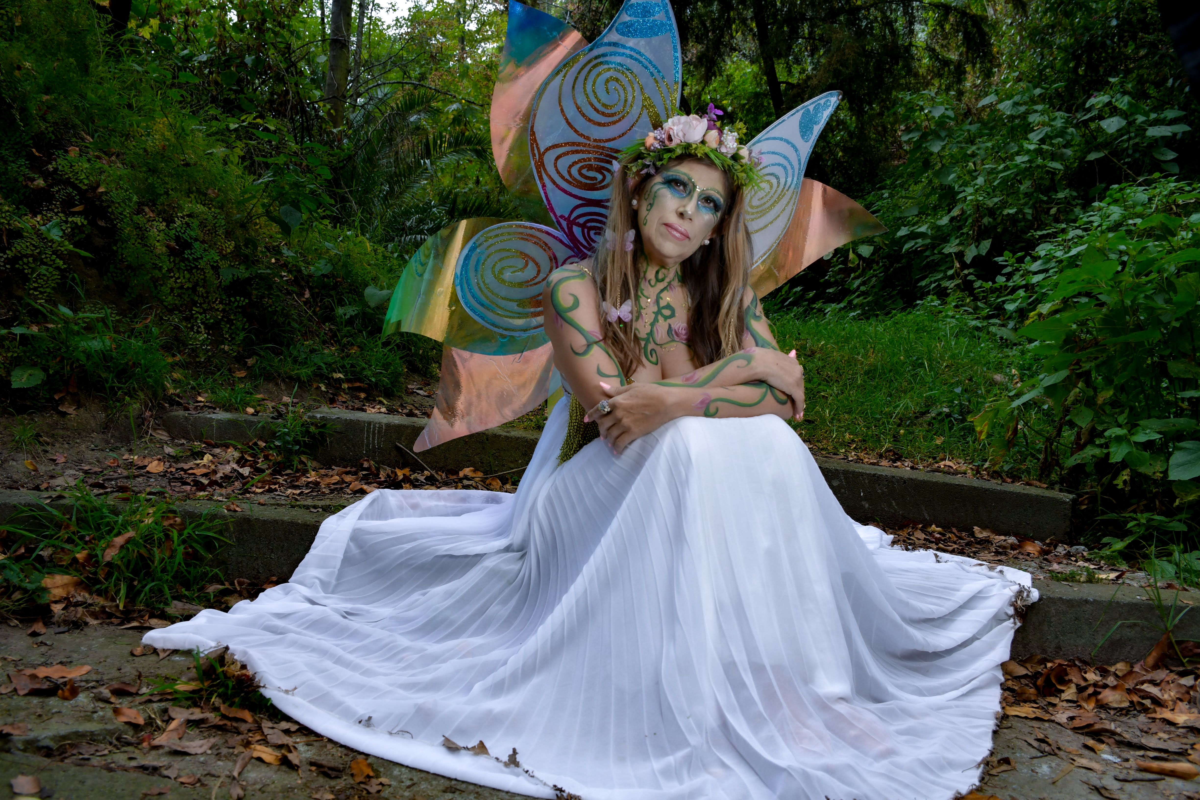 Fairy woman in a white gown with colorful wings sitting in a lush forest.