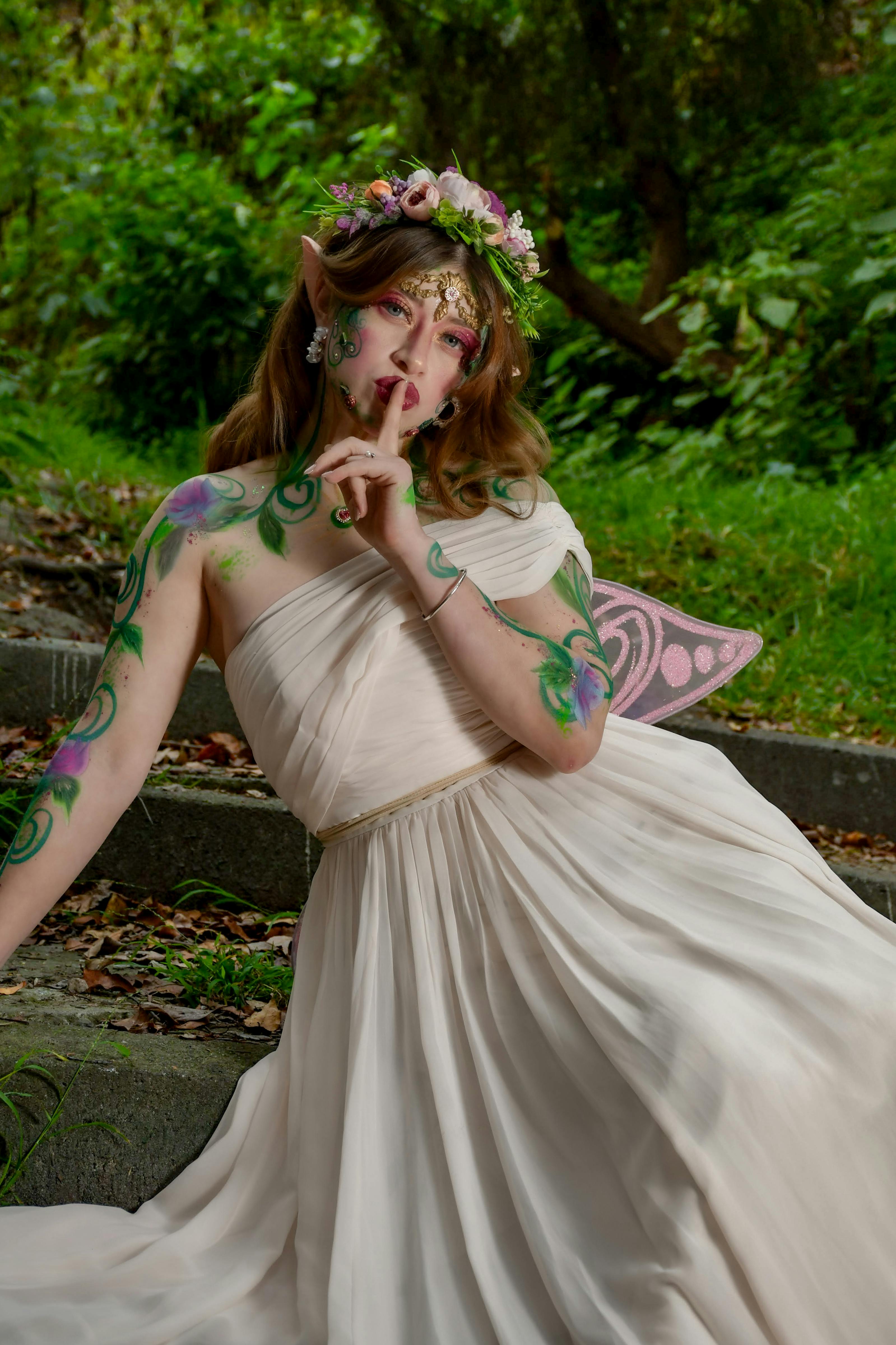 A whimsical fairy in a flowing white gown with floral headpiece sits in a lush green forest.