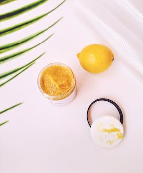 Aesthetic flat lay of lemon scrub jar beside a lemon and palm leaves on a white surface.
