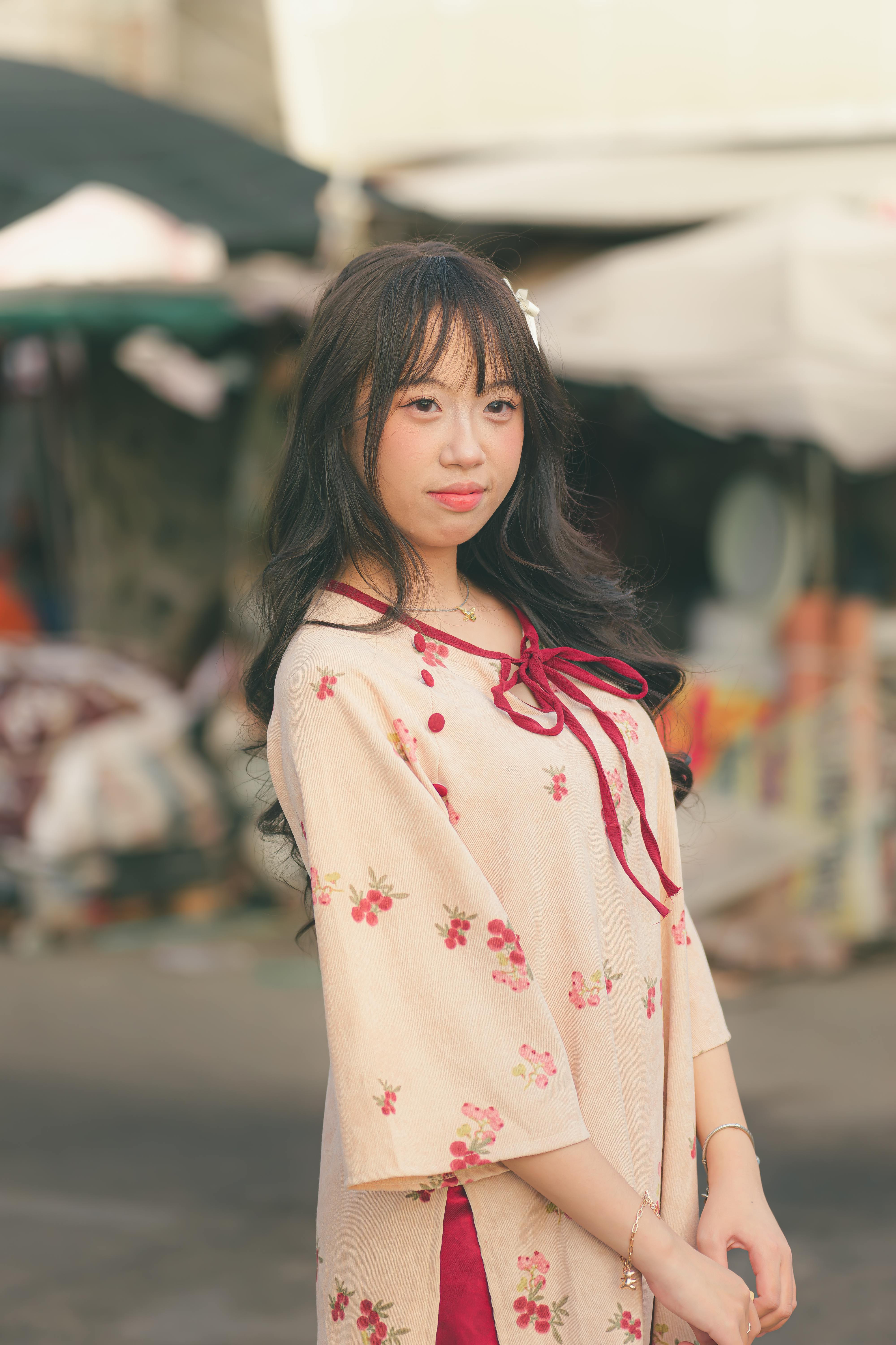 A young woman wearing a floral traditional dress poses outdoors in soft lighting.