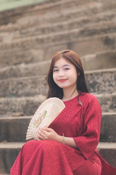 Young woman in red traditional dress sitting on steps holding a fan.