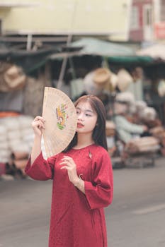 A woman in a red dress holding a fan at a bustling outdoor market.
