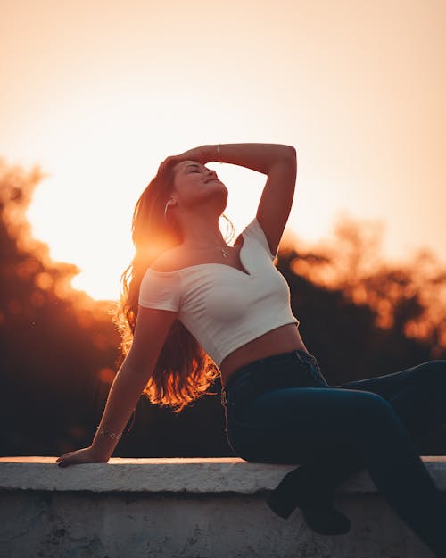 Foto Wanita Tersenyum Duduk Di Pagar Batu Berpose Dengan Mata Tertutup Saat Golden Hour