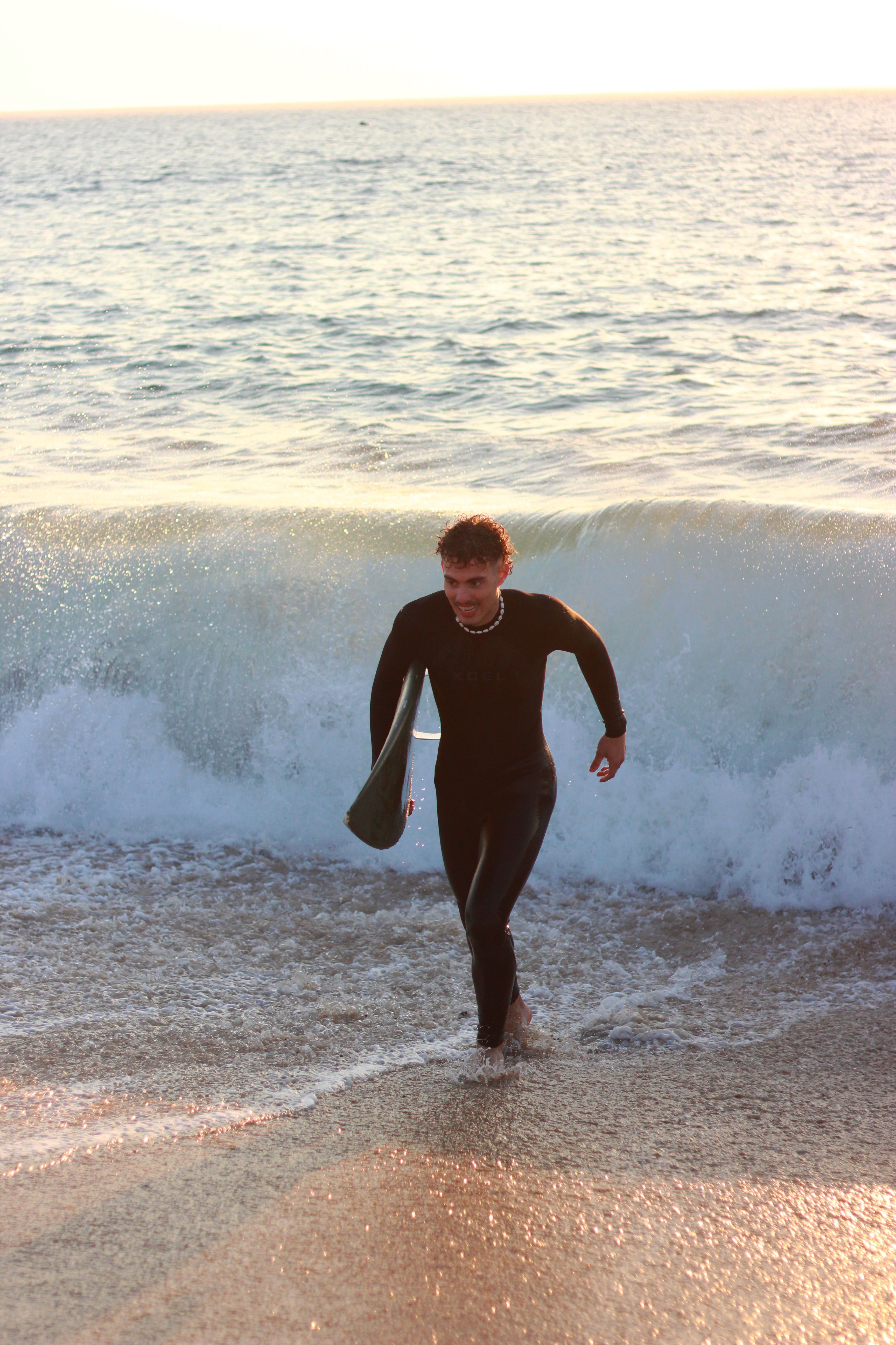 A surfer wearing a wetsuit emerges from the ocean at Nazaré Beach, Portugal.
