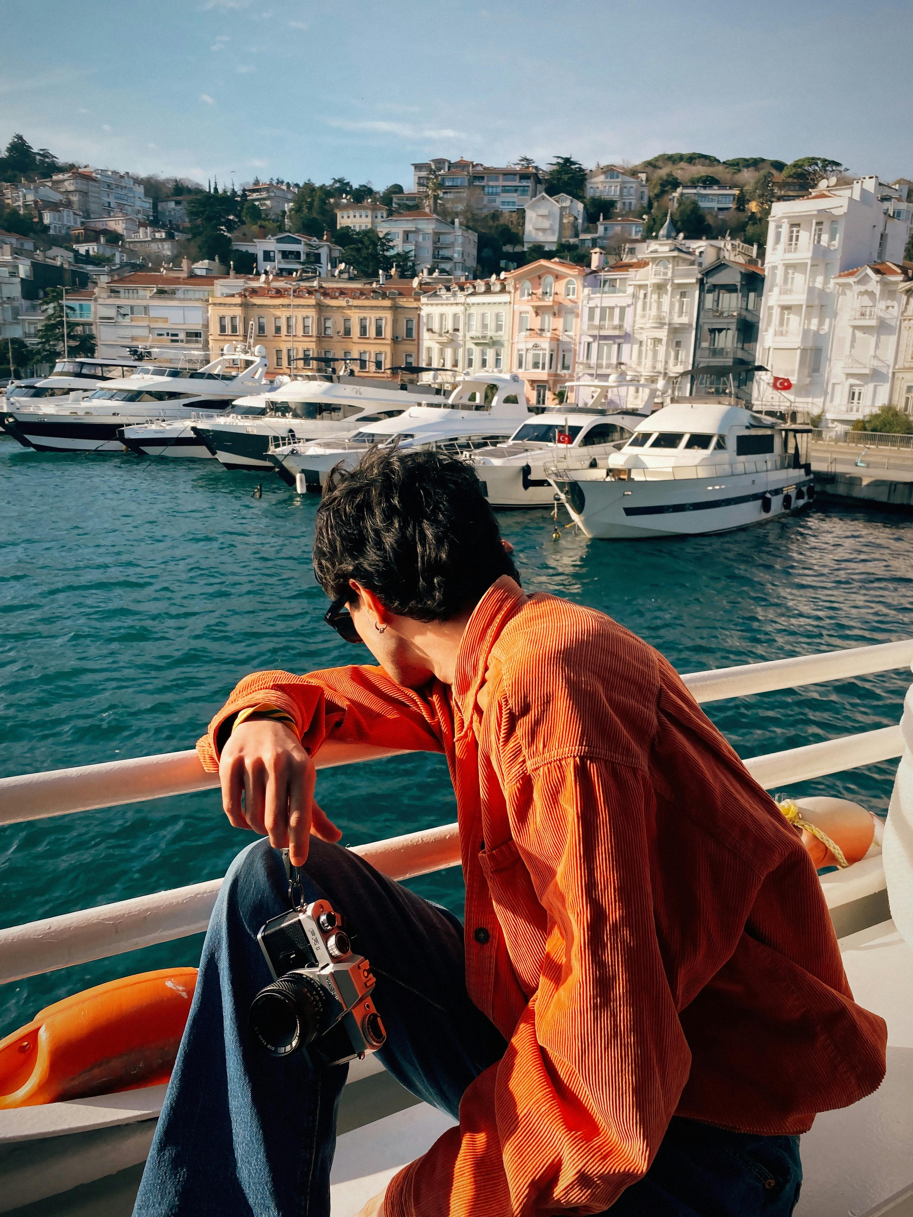 Person seated on a boat, looking at a scenic harbor view with yachts and colorful buildings.