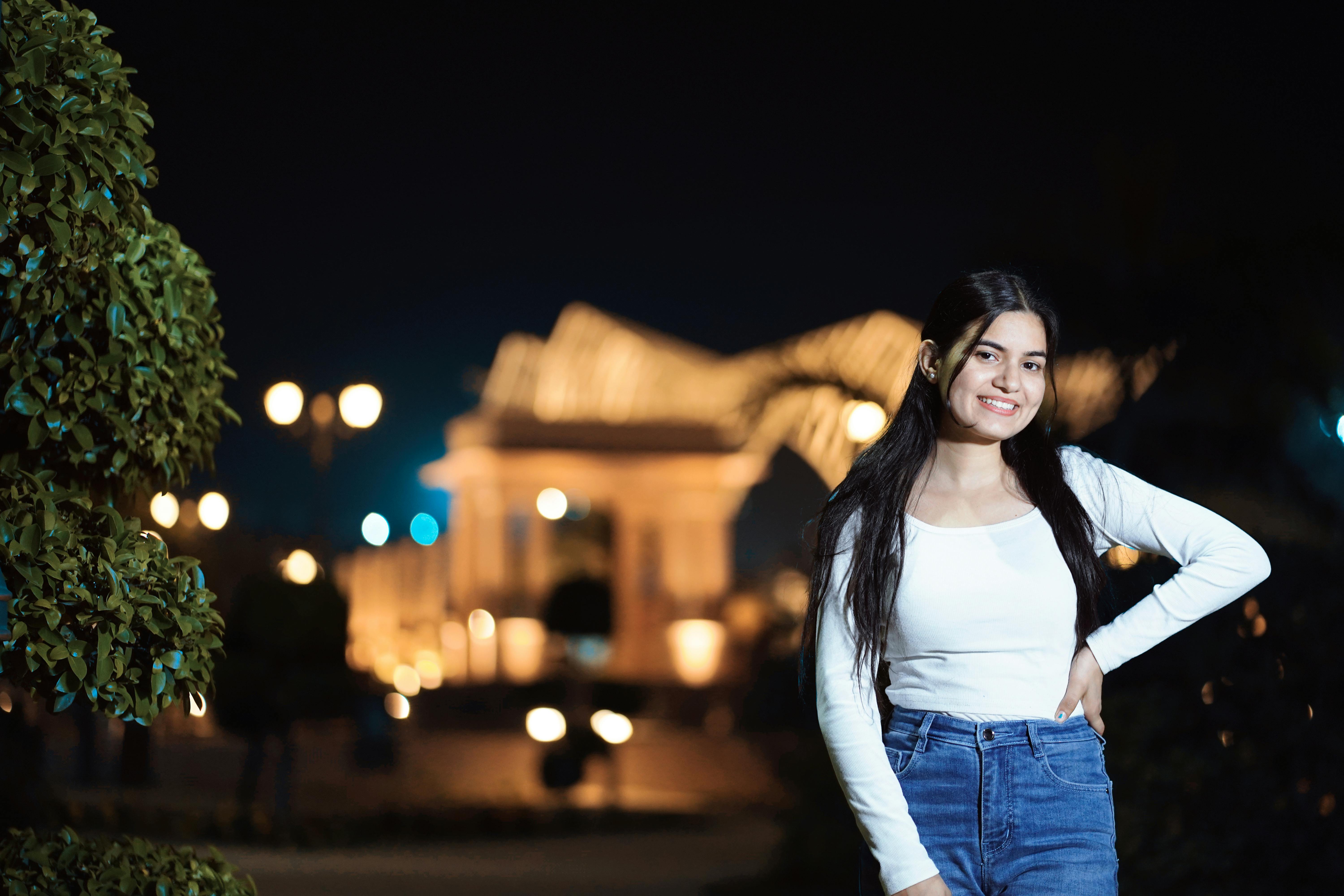 A young woman poses confidently outdoors at night with a lit building in the background.