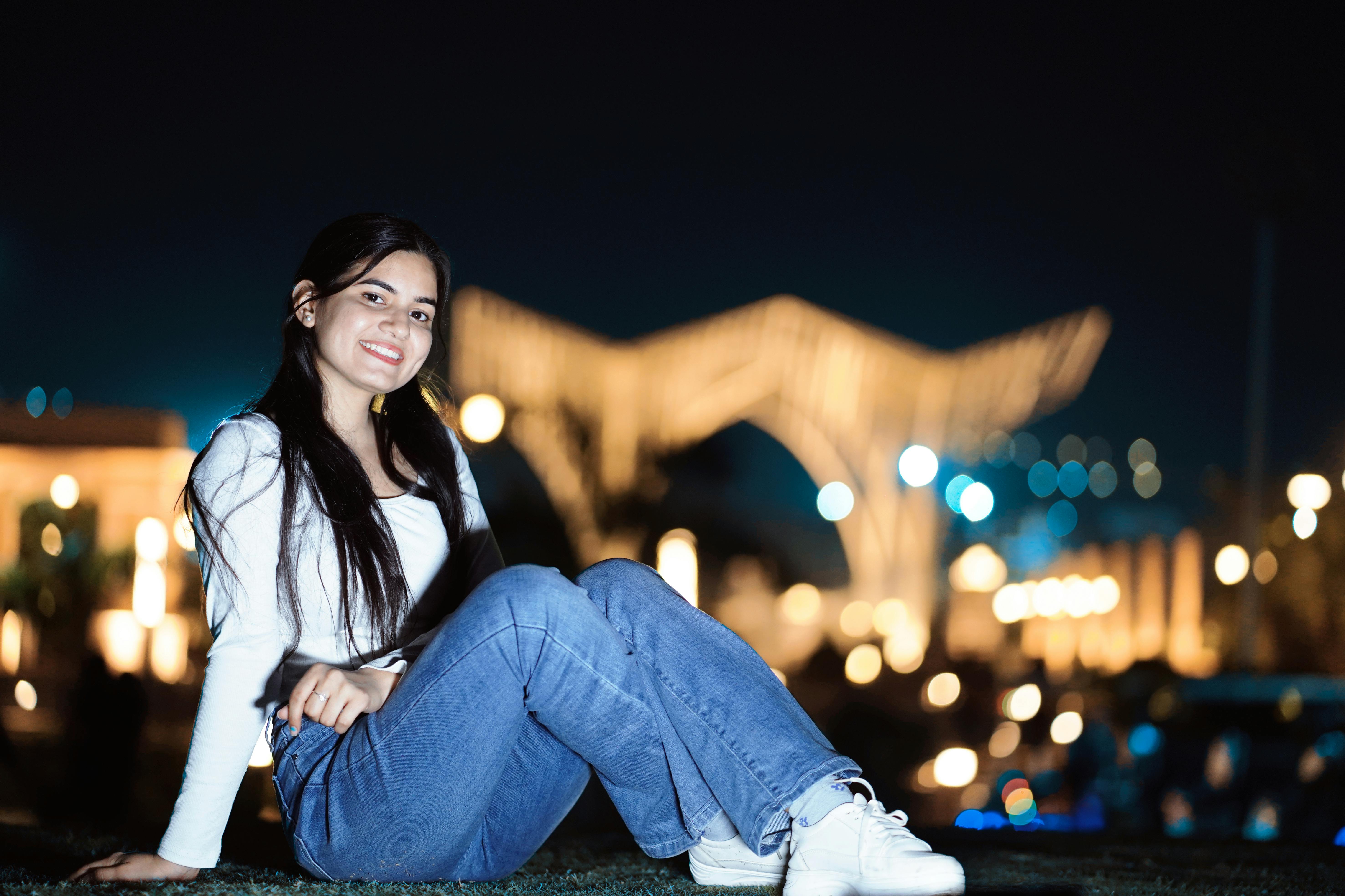 Free Young woman posing with a smile in an outdoor nighttime setting with blurred lights. Stock Photo