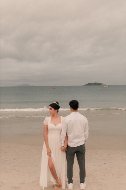 Free A couple in wedding attire holds hands on a serene beach with cloudy skies. Stock Photo