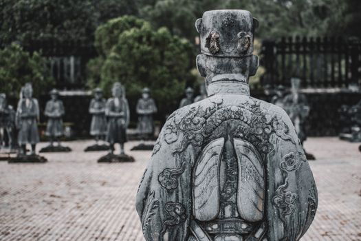 Back view of an intricately carved stone warrior statue among others in a serene outdoor space.