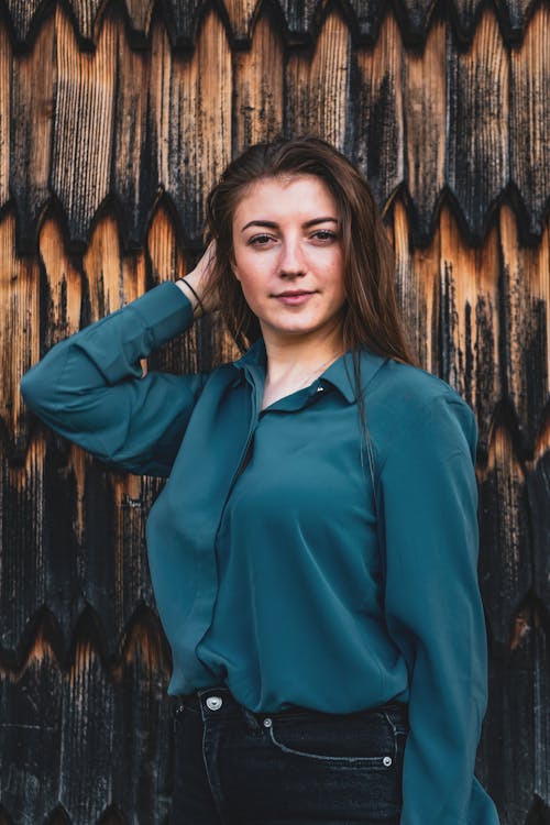 Photo of Woman in Blue Button-up Blouse and Black Jeans Posing In Front of Wooden Wall