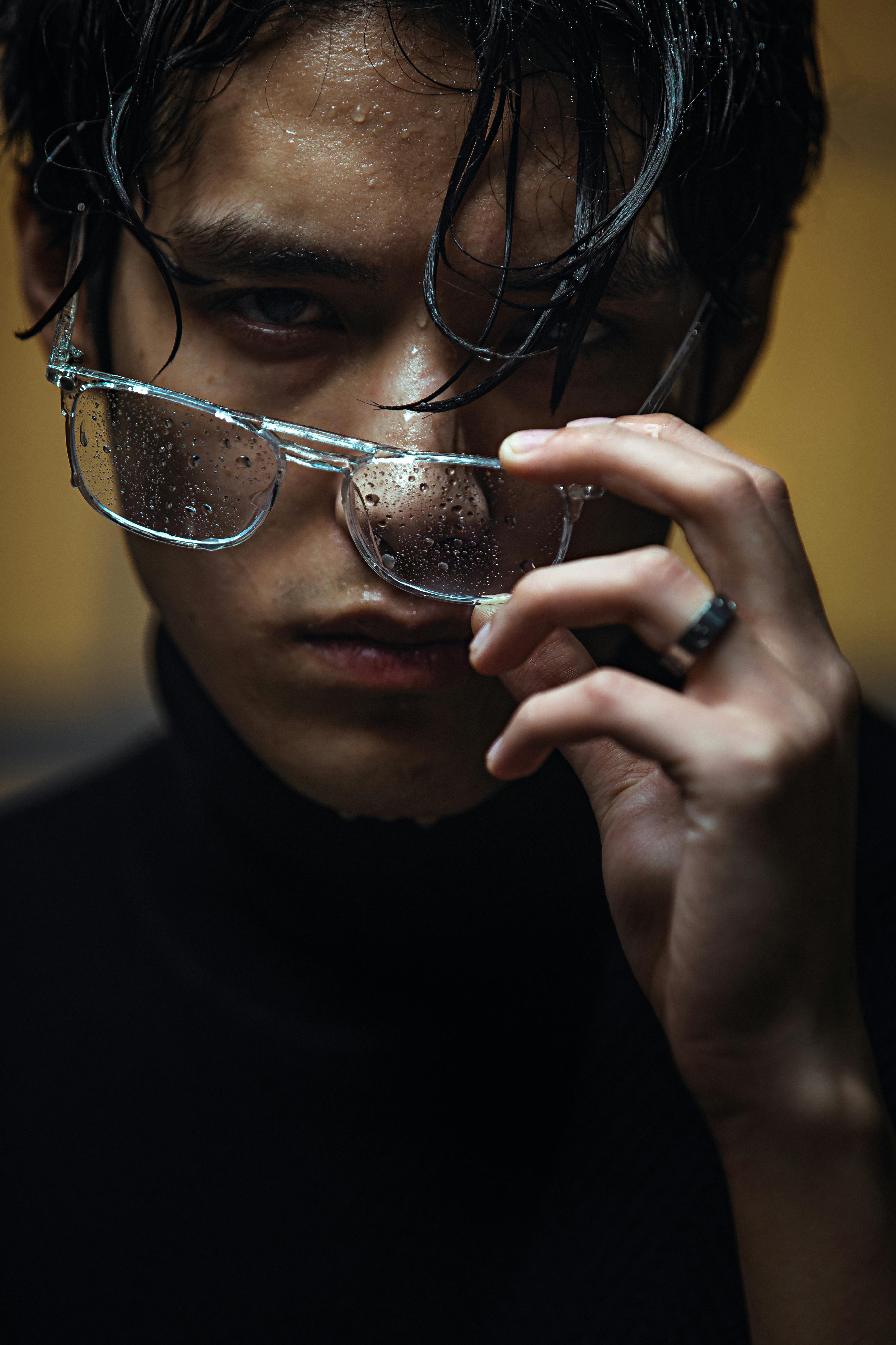 Close-up of a man with wet glasses adjusting them, creating a moody and dramatic fashion look.