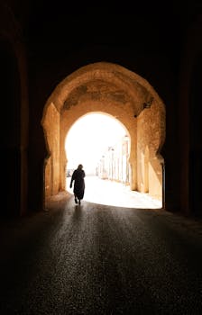 A silhouette of a person walking through a traditional Moroccan tunnel with sunlight streaming in.