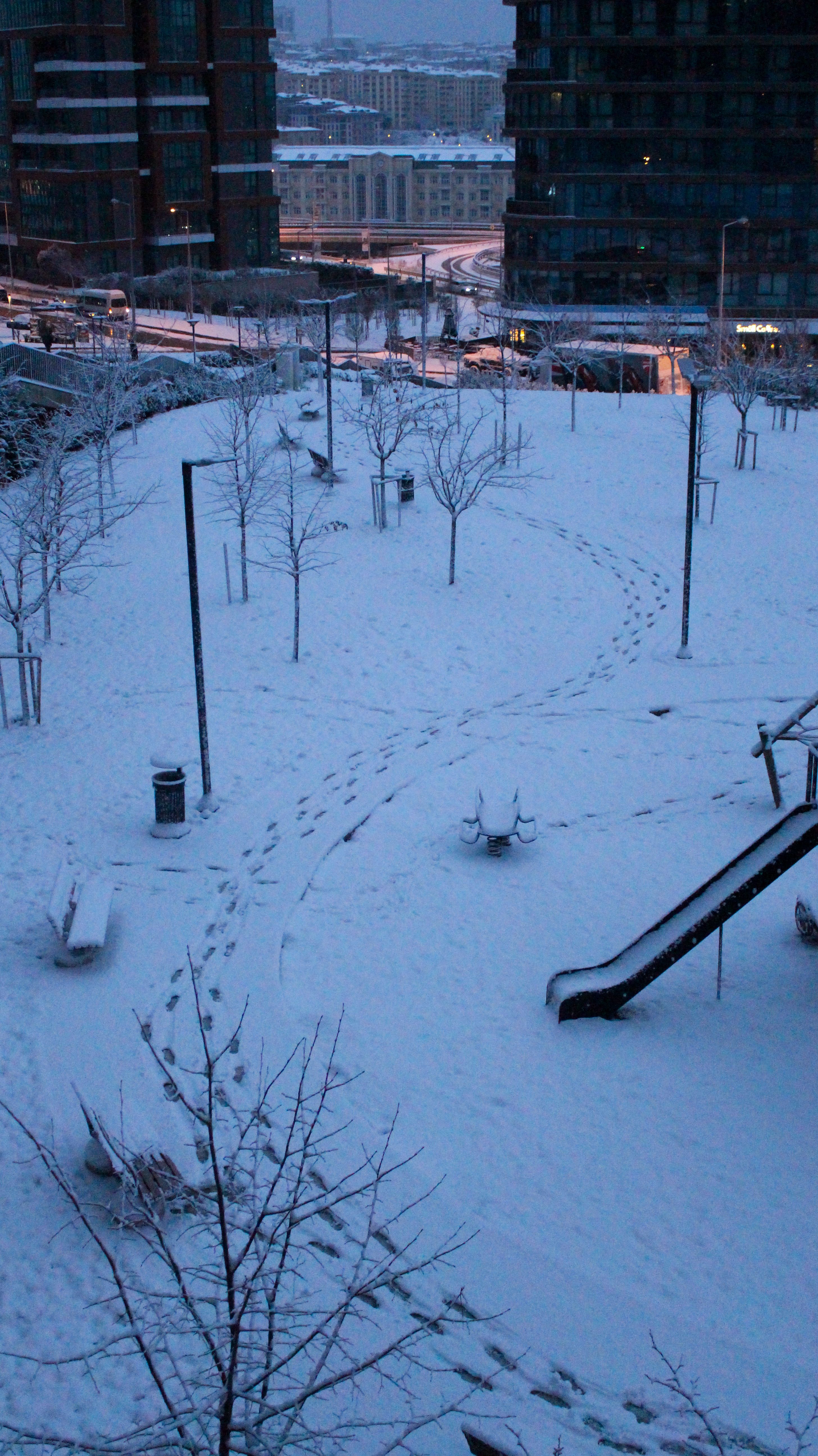 A serene urban park covered in snow during a winter evening with visible footprints.