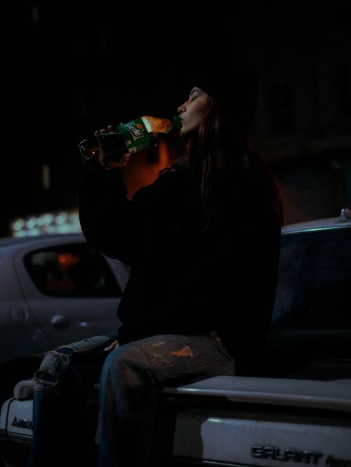 Free Woman drinking a bottle of soda outdoors at night, sitting on a car, captured with a moody ambiance. Stock Photo
