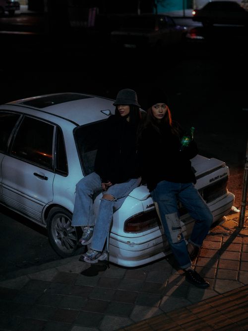 Free Two individuals in casual attire sitting on a parked car at night in an urban setting. Stock Photo