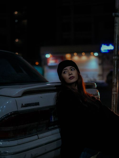 Free Dramatic nighttime portrait of a woman in a beanie leaning on a car. Stock Photo