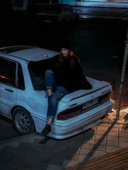 Mysterious night shot of a woman sitting on a car, casting moody urban vibes.