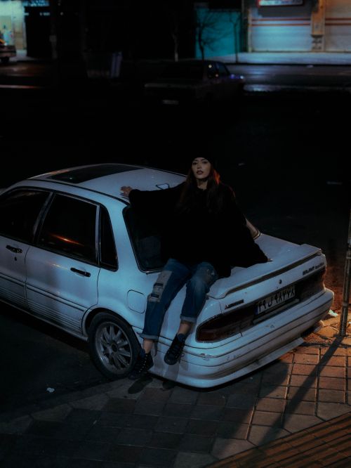 Free A young woman in a beanie sits on a car trunk under street lamps at night. Stock Photo