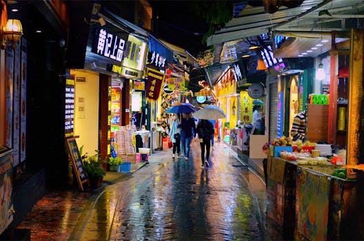 Busy night market street with colorful shops and people holding umbrellas.