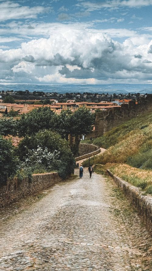 Free Two people walking down a cobblestone path in a historic medieval village, surrounded by lush greenery. Stock Photo