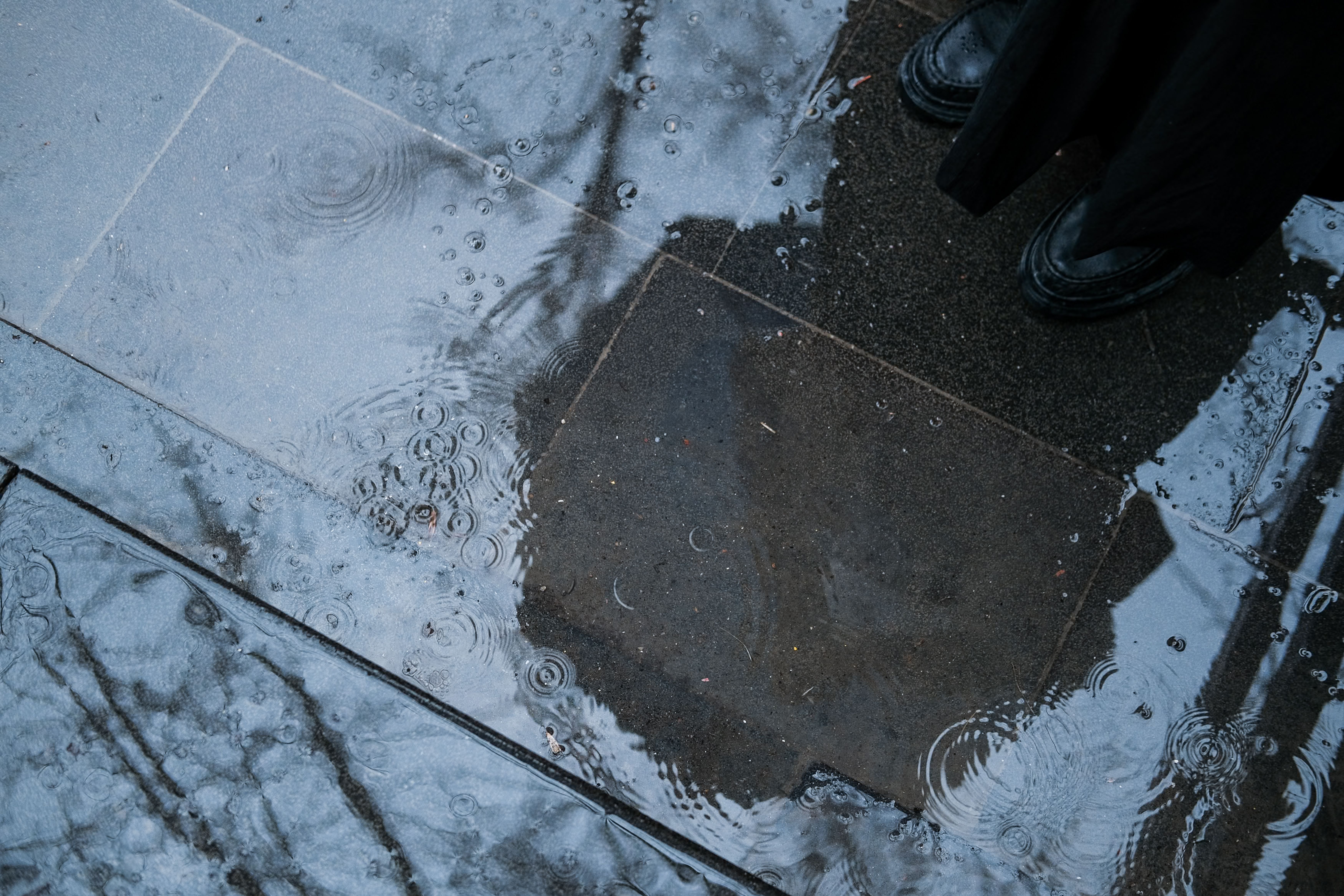 View of wet sidewalk with reflective rain puddles and a person's feet in boots.