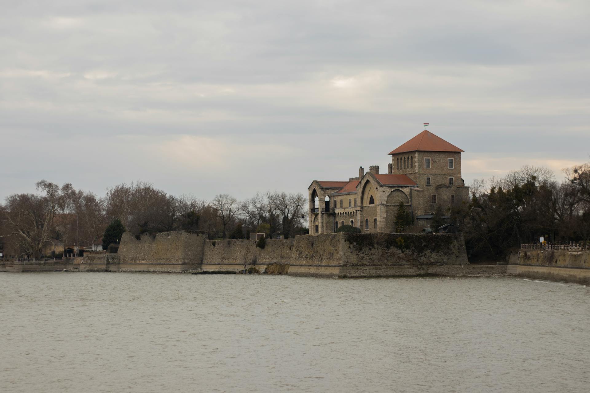 A serene view of a historic castle beside a calm lake reflecting overcast skies.