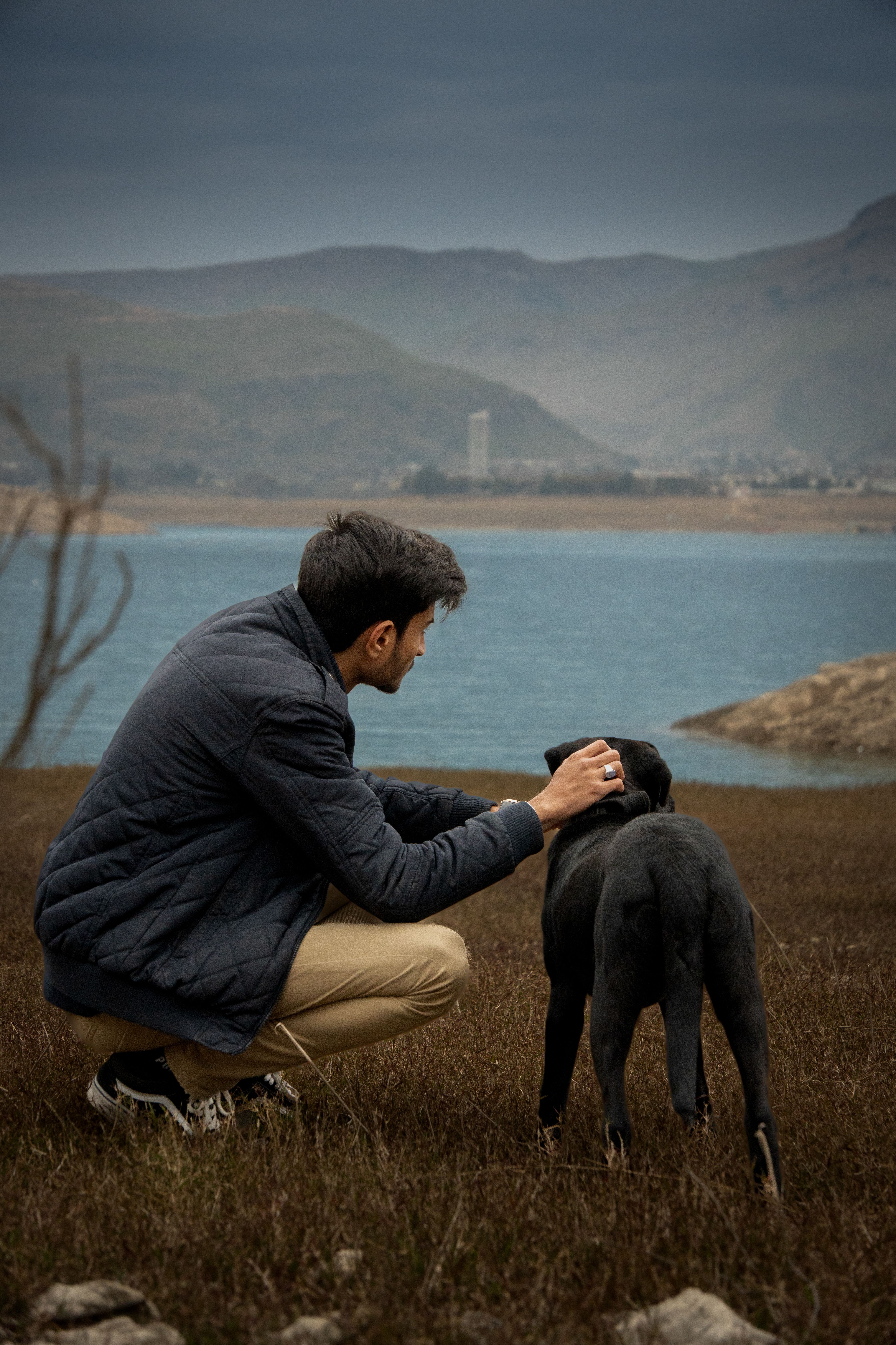 A man in a jacket kneeling besides a black dog overlooking a serene lake.