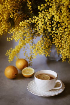 A warm cup of tea with lemon slices by vibrant yellow mimosa flowers.