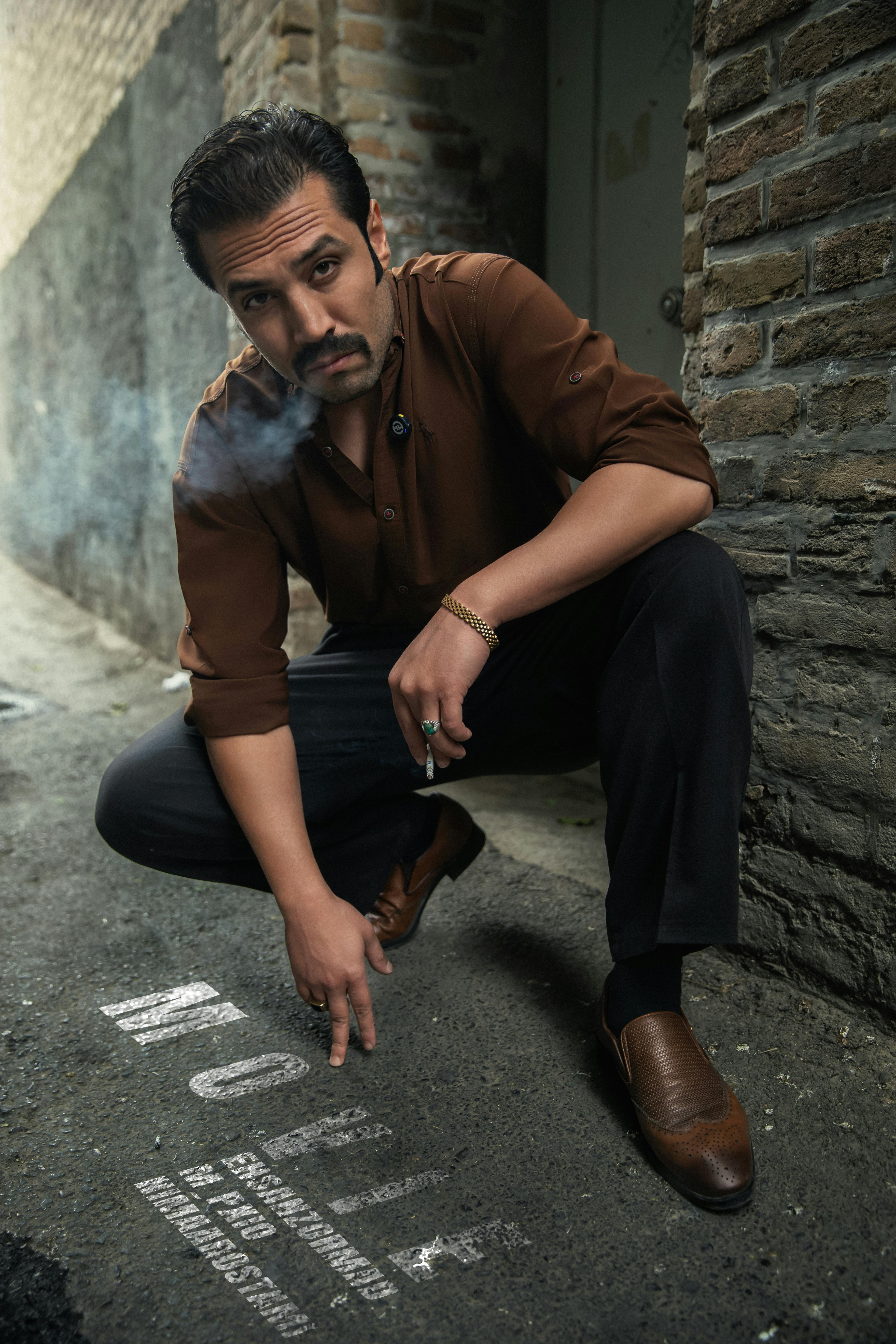 Man squatting in a narrow alley, smoking a cigarette and pointing at graffiti on the ground.