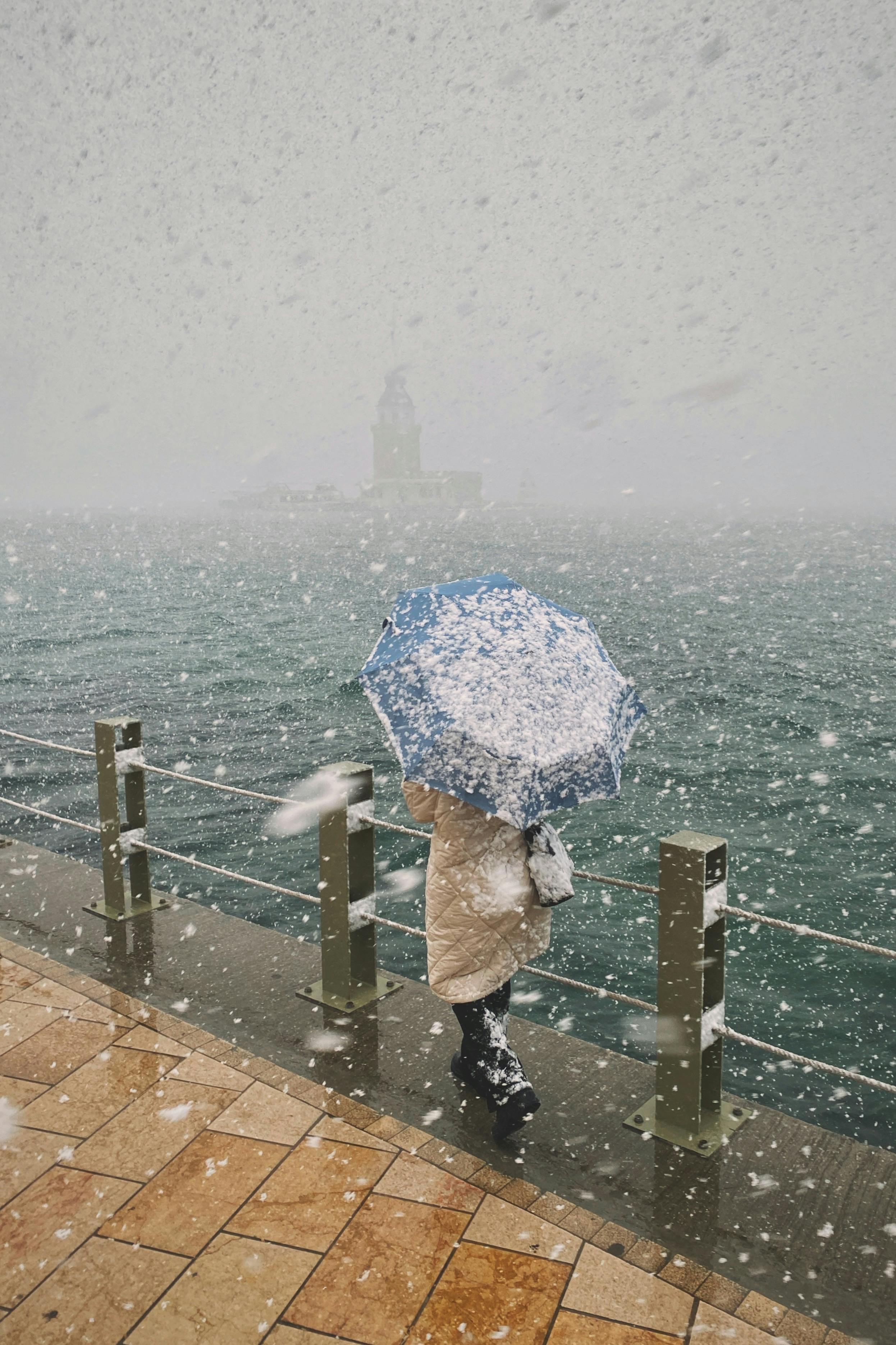 一个人拿着一把蓝色的雨伞，走在白雪皑皑的海滨上