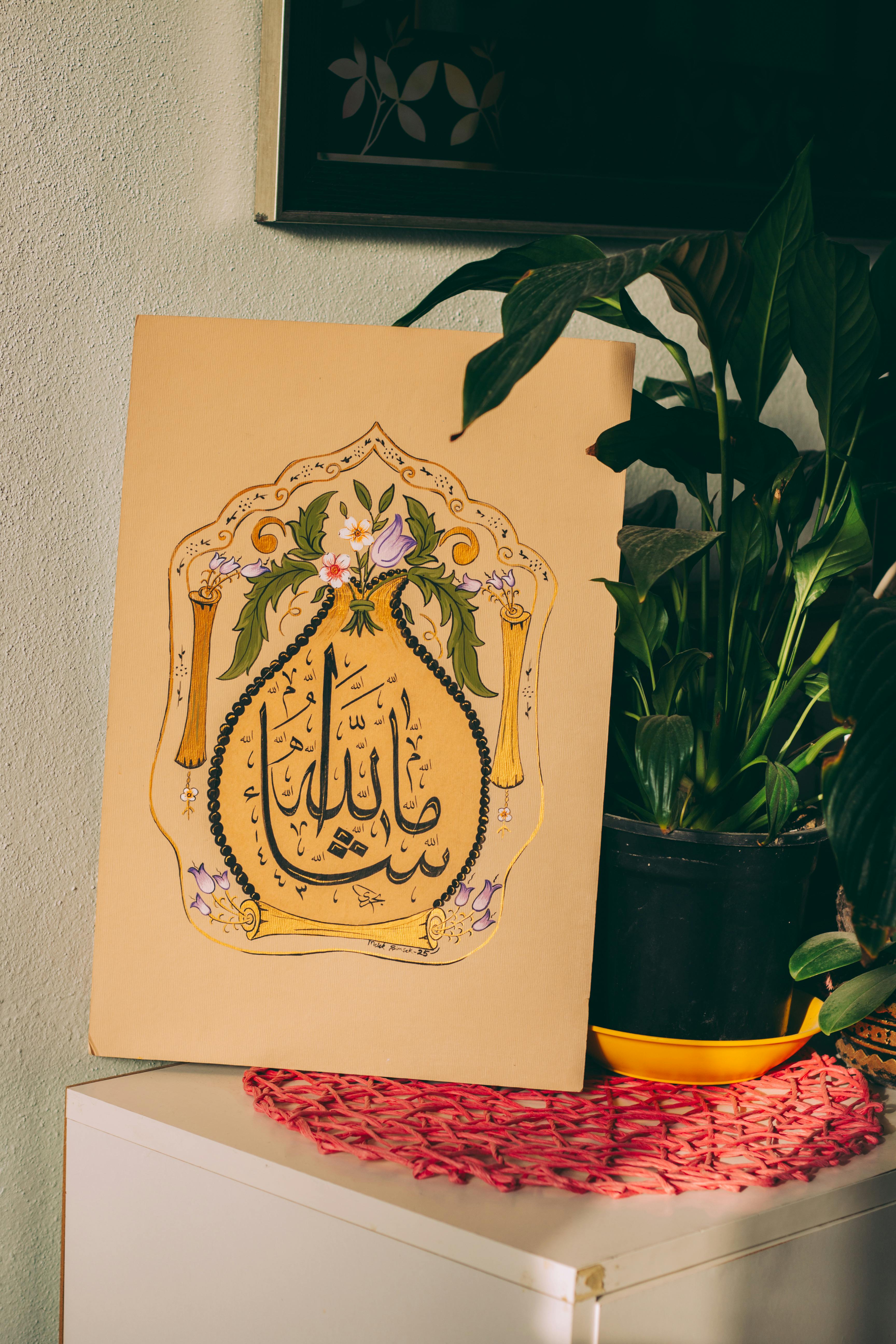 Artistic Arabic calligraphy with floral elements next to a houseplant on a shelf.